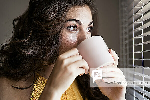 Nahaufnahme einer jungen Frau  die an einer Tasse Kaffee nippt  während sie durch die Jalousien ihres Hauses schaut