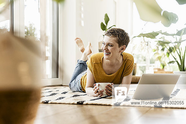 Fröhliche Frau schaut weg  während sie mit Kaffeetasse und Laptop auf dem Teppich zu Hause liegt
