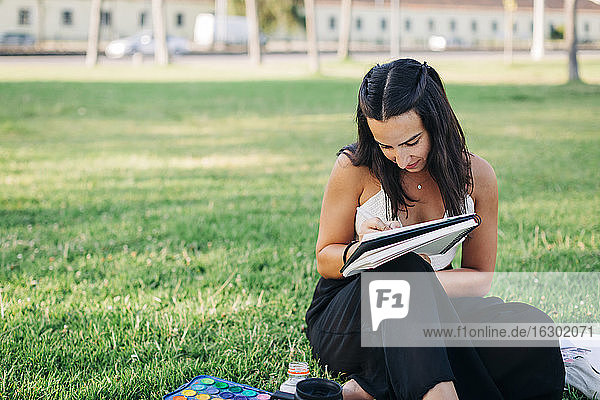Woman painting in book while sitting on grass in public park