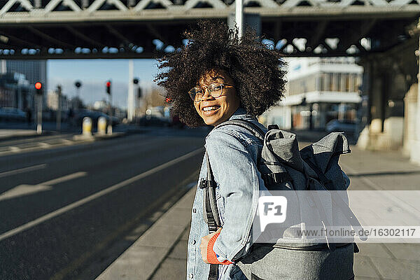 Lächelnde Afro-Frau mit Rucksack steht auf dem Bürgersteig in der Stadt an einem sonnigen Tag