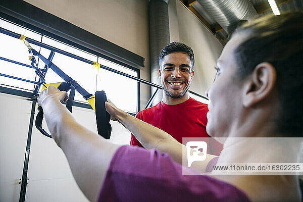 Lächelnder Fitnesstrainer  der einer Frau beim Training mit Riemen im Fitnessstudio hilft