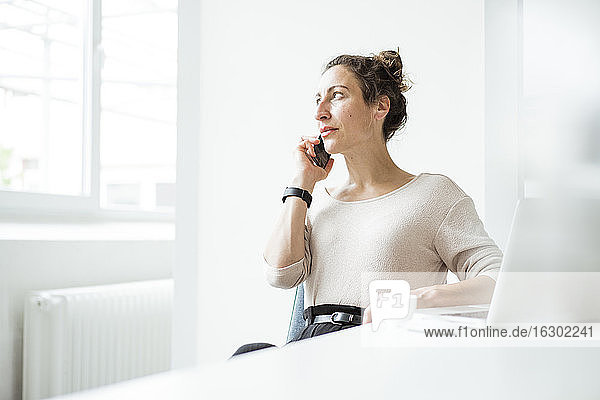 Businesswoman talking over smart phone while sitting with laptop on table