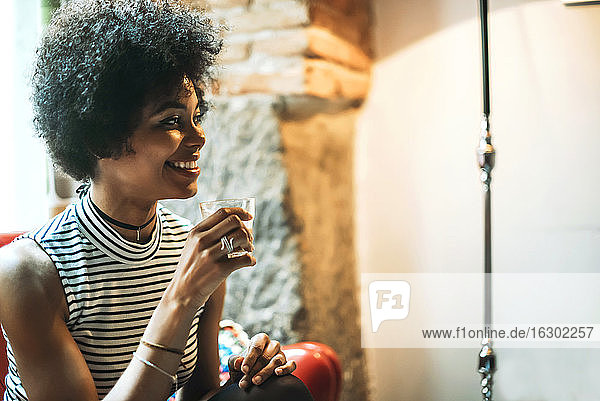 Nahaufnahme einer lächelnden Frau mit lockigem Haar  die einen Kaffee in der Hand hält und an der Wand eines Cafés sitzt