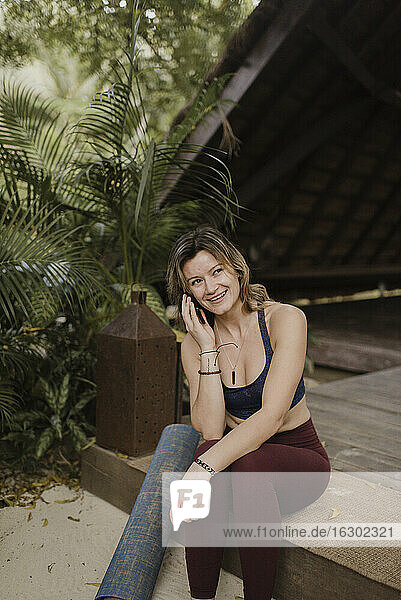 Lächelnde Frau mit Yogamatte und Smartphone in einer Hütte