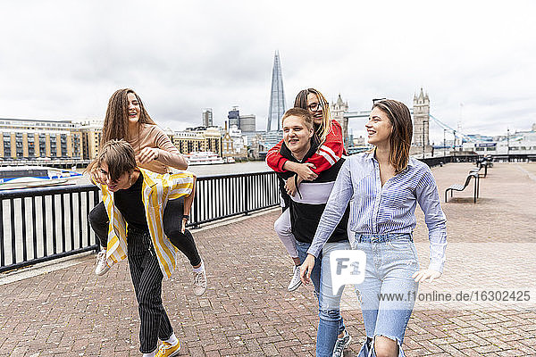 Männliche Freunde  die ihre Freundinnen beim Gehen auf einem Fußweg in der Stadt London huckepack nehmen  Großbritannien