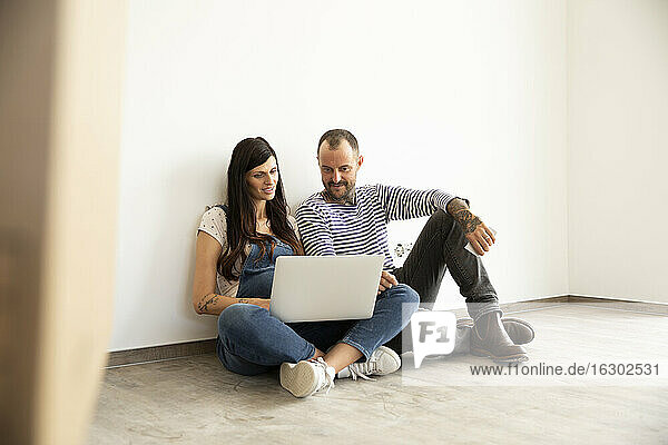 Ein Paar benutzt einen Laptop und sitzt an einer weißen Wand in einer neuen  unmöblierten Wohnung