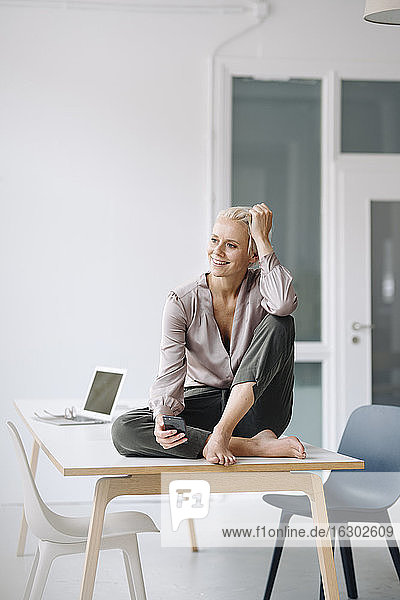 Nachdenkliche Unternehmerin  die ihr Smartphone in der Hand hält  während sie auf dem Schreibtisch an der Wand im Büro sitzt