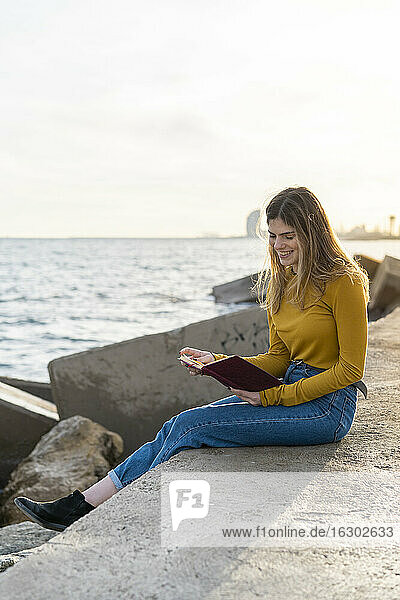 Junge Frau mit Tagebuch sitzend am Meer gegen den Himmel