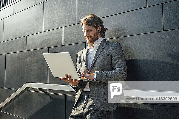 Gut aussehender junger Mann  der einen Laptop benutzt  während er in der Innenstadt an der Wand steht