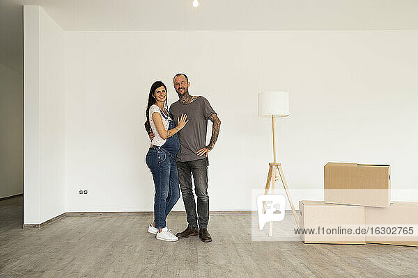 Smiling couple with electric lamp and boxes standing against wall in new house
