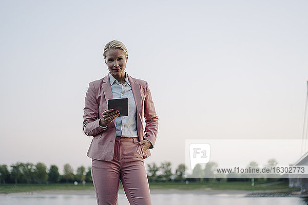Selbstbewusste Geschäftsfrau  die ein digitales Tablet hält  während sie mit der Hand in der Tasche vor einem klaren Himmel bei Sonnenuntergang steht