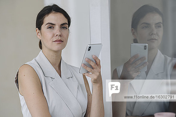 Confident businesswoman using mobile phone while standing by glass wall at office