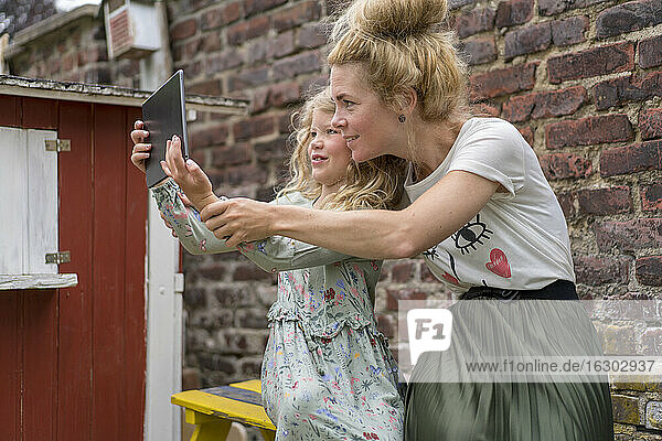 Smiling girl taking selfie with mother through digital tablet against brick wall at back yard