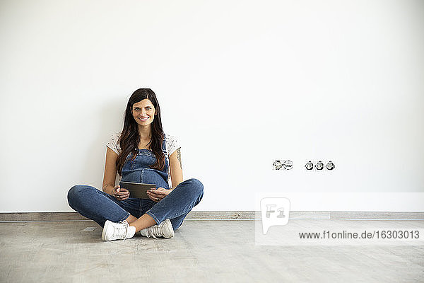 Lächelnde schwangere Frau mit digitalem Tablet  die auf dem Boden vor einer weißen Wand in einem neuen Haus sitzt