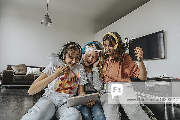 Cheerful friends listening music through headphones while using digital tablet at home