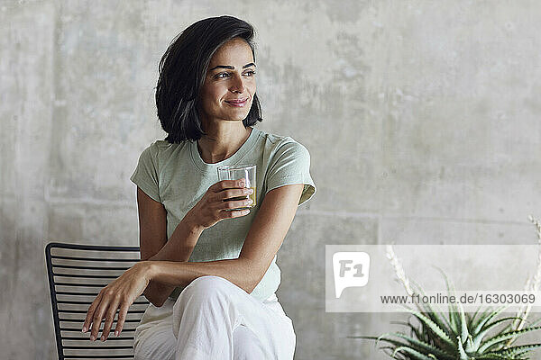 Smiling businesswoman holding smoothie while sitting on chair against wall in office