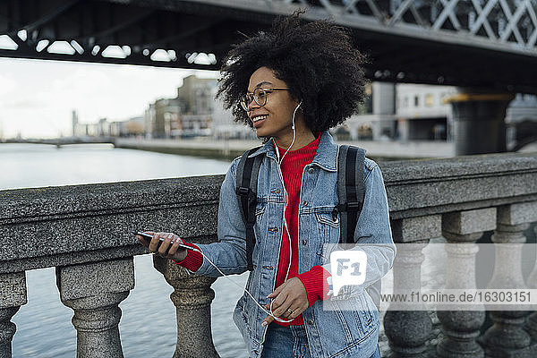Junge Afroamerikanerin hört Musik über Kopfhörer  während sie am Geländer einer Fußgängerbrücke steht