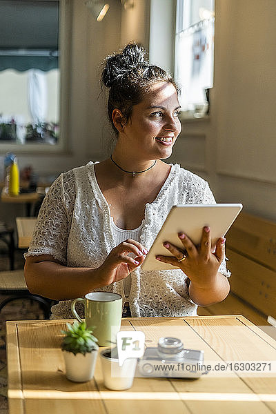 Lächelnde üppige Frau  die ein digitales Tablet benutzt und wegschaut  während sie an einem Tisch im Café sitzt