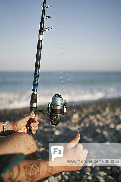 Close-up of mid adult man holding fishing rod at beach against clear sky during sunset