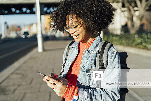 Nahaufnahme einer lächelnden jungen Frau mit lockigem Haar  die ein Mobiltelefon benutzt  während sie auf dem Gehweg steht