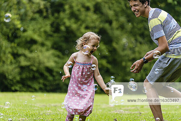 Fröhlicher Bruder spielt mit Schwester inmitten von Seifenblasen im öffentlichen Park