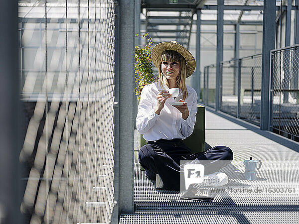 Geschäftsfrau mit Hut und Kaffeetasse am Zaun im Gewächshaus sitzend