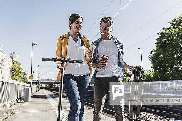 Lächelnde Frau  die einem männlichen Freund beim Spaziergang am Bahnhof ihr Smartphone zeigt