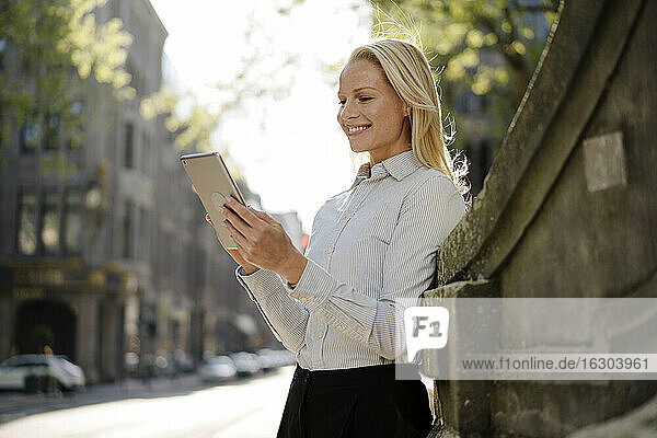 Smiling blond female professional using digital tablet while leaning on surrounding wall in city
