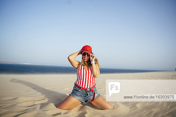 Cheerful woman holding cap while kneeling on sand at beach against clear blue sky