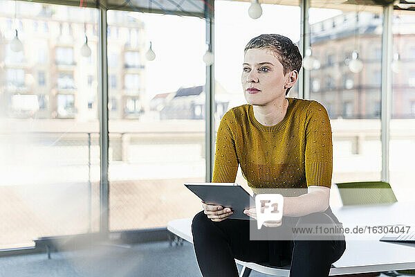 Nachdenkliche Geschäftsfrau sitzt auf dem Schreibtisch im Büro und hält ein Tablet