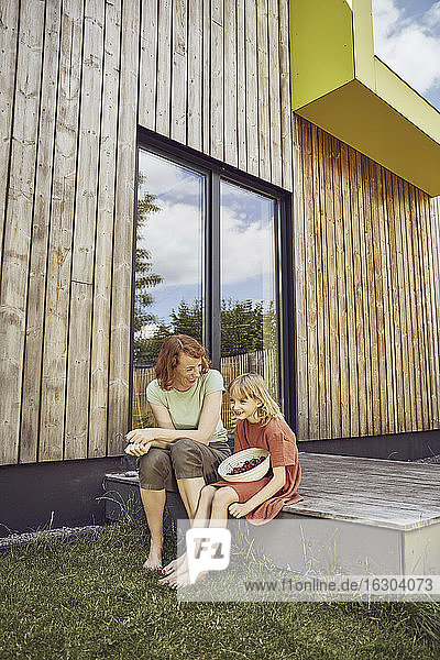 Mutter und Tochter mit Kirschen vor einem kleinen Haus sitzend