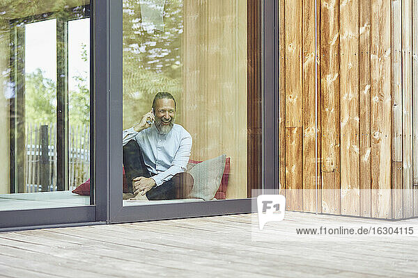 Cheerful man talking over smart phone while sitting on bed in tiny house seen through window