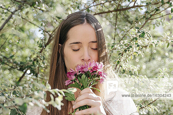 Frau riecht an frischen rosa Blumen,  während sie im Frühling in einem Park unter Bäumen steht