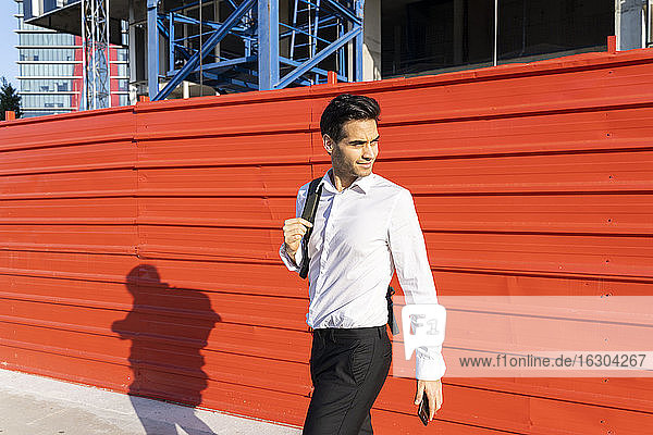 Geschäftsmann schaut weg  während er an einer roten Mauer in der Stadt an einem sonnigen Tag spazieren geht