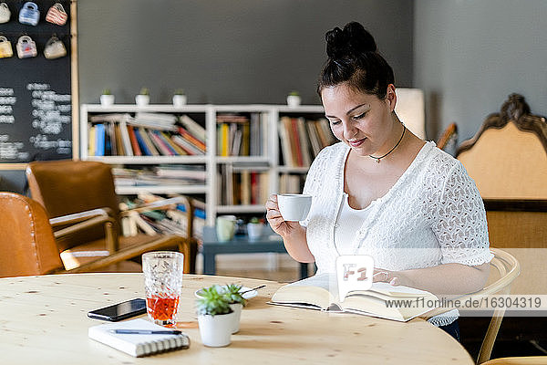 Üppige Frau hält eine Kaffeetasse und liest ein Buch auf einem Tisch im Restaurant