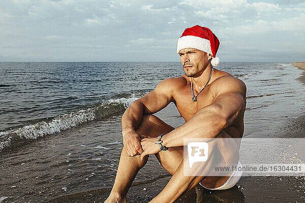 Nachdenklicher junger Mann ohne Hemd mit Weihnachtsmannmütze am Strand sitzend