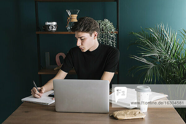 Gut aussehender junger Mann schreibt in ein Buch  während er mit einem Laptop am Tisch sitzt