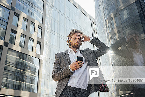Besorgter männlicher Berufstätiger mit Kopf in der Hand  der sein Smartphone benutzt  während er sich bei der Reflexion über ein Bürogebäude in der Innenstadt anlehnt