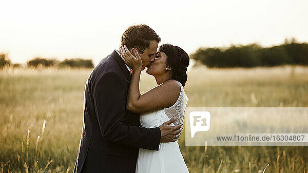 Affectionate young bridal couple kissing at field during sunset