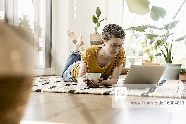 Mittlere erwachsene Frau  die eine Kaffeetasse hält und einen Laptop benutzt  während sie zu Hause auf dem Teppich liegt