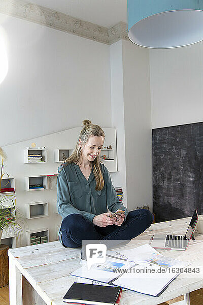 Lächelnde Frau sitzt auf dem Schreibtisch im Heimbüro und benutzt ein Smartphone