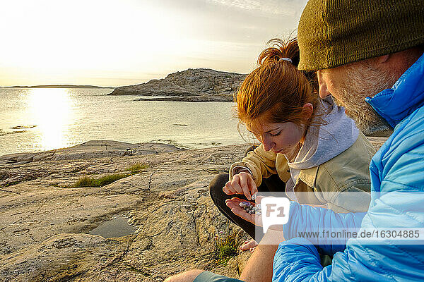 Schweden  Bezirk Vastra Gotaland  Grebbestad  Mann und junges Mädchen sammeln Kieselsteine am felsigen Ufer des Tjurpannans Naturreservats bei Sonnenuntergang