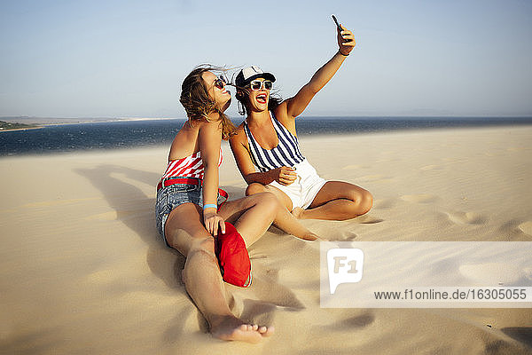 Cheerful woman taking selfie with friend while sitting on sand at beach against clear sky