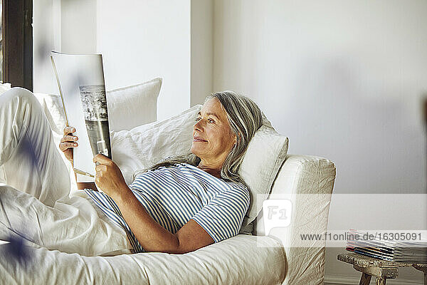 Frau liest eine Zeitschrift  während sie auf dem Sofa im Wohnzimmer liegt