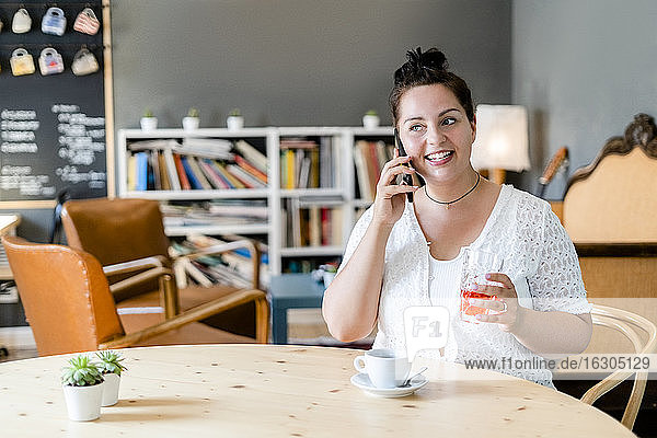 Üppige Frau  die ein Getränk hält und über ein Smartphone spricht  während sie in einem Café sitzt