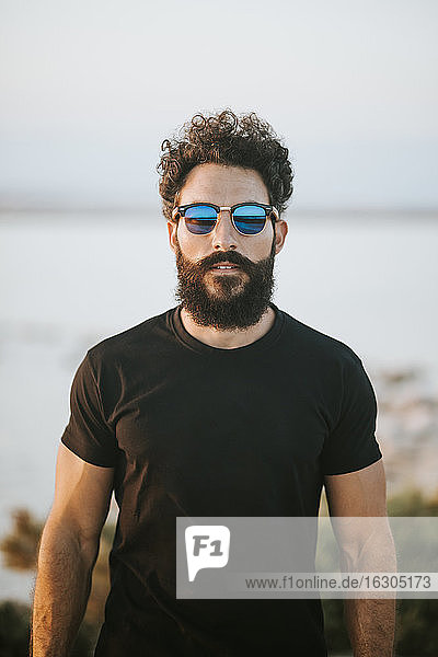 Bearded man wearing sunglasses while standing against lake