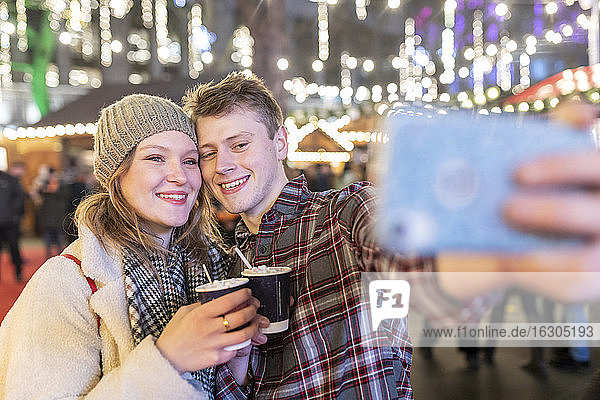 Paar hält heiße Schokolade und macht ein Selfie auf dem nächtlichen Weihnachtsmarkt