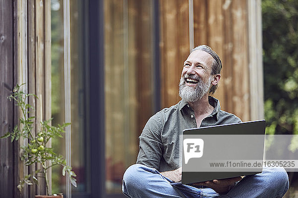 Fröhlicher reifer Mann mit Laptop  der wegschaut  während er vor einem kleinen Haus sitzt