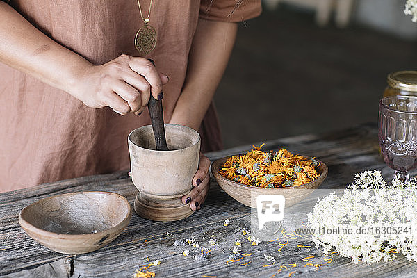 Midsection of woman crushing flowers and herbs in mortar with pestle on wooden table