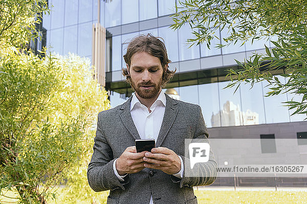Hübscher Geschäftsmann mit Bart  der ein Smartphone benutzt  während er vor einem Bürogebäude steht